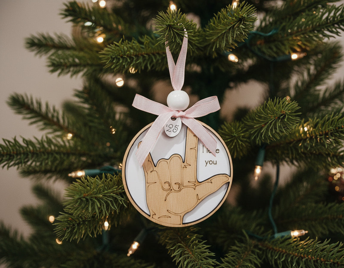 ASL ornament with a hand sign for 'I love you' and a pink ribbon, pale hand on a white background. Accented with the current year of 2025 charm and wood bead.