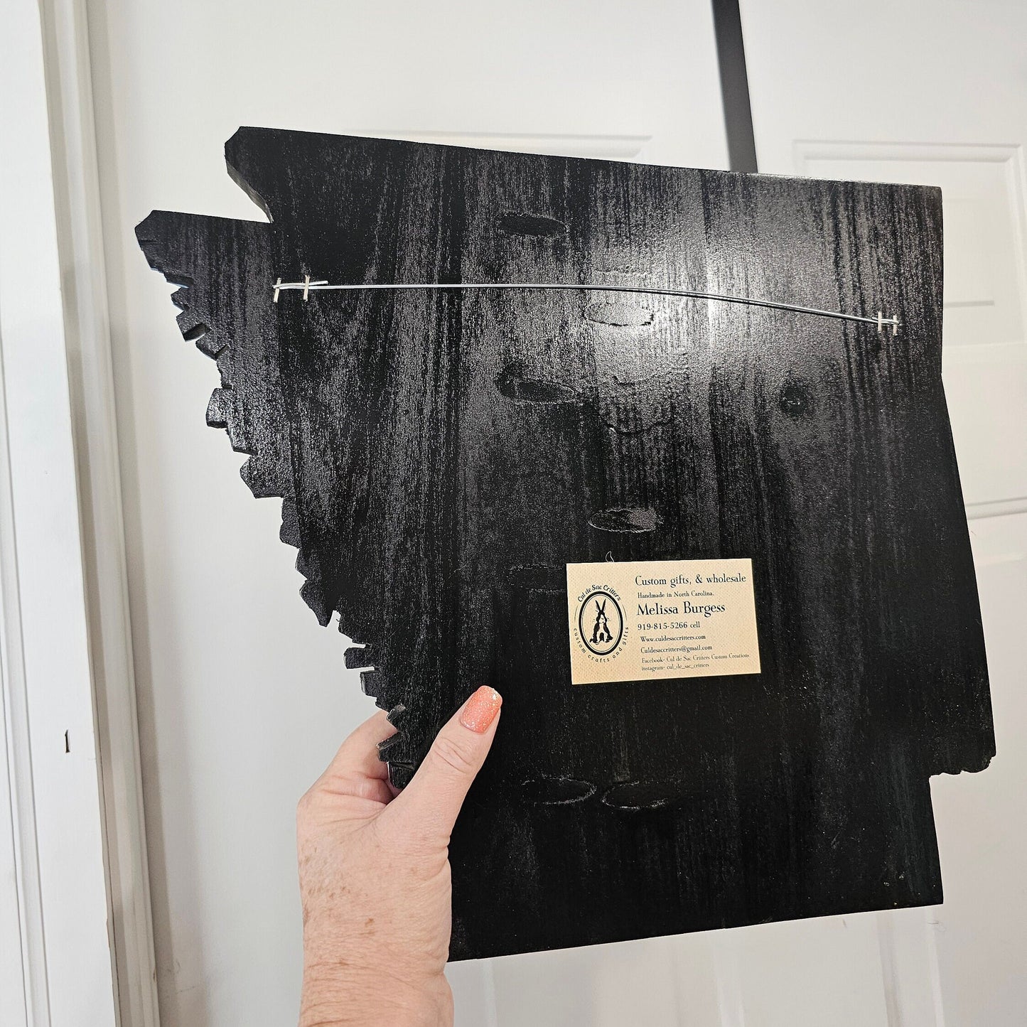Black wooden back of Arkansas wall art shaped like a state, held by a hand against a white door.