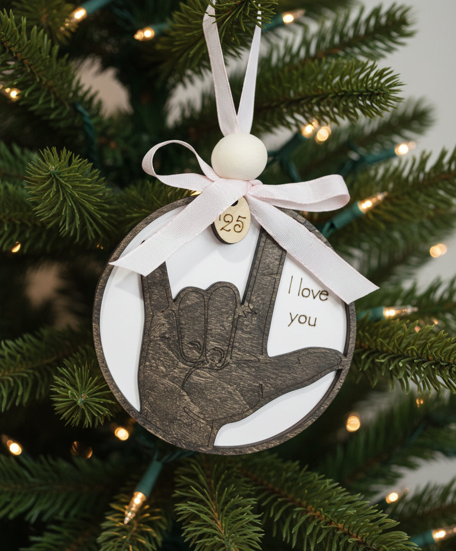 Dark hand with ASL sign, 'I love you' on a white background, hand-tied pale pink check ribbon, current year charm of 2025 and wood bead accent.