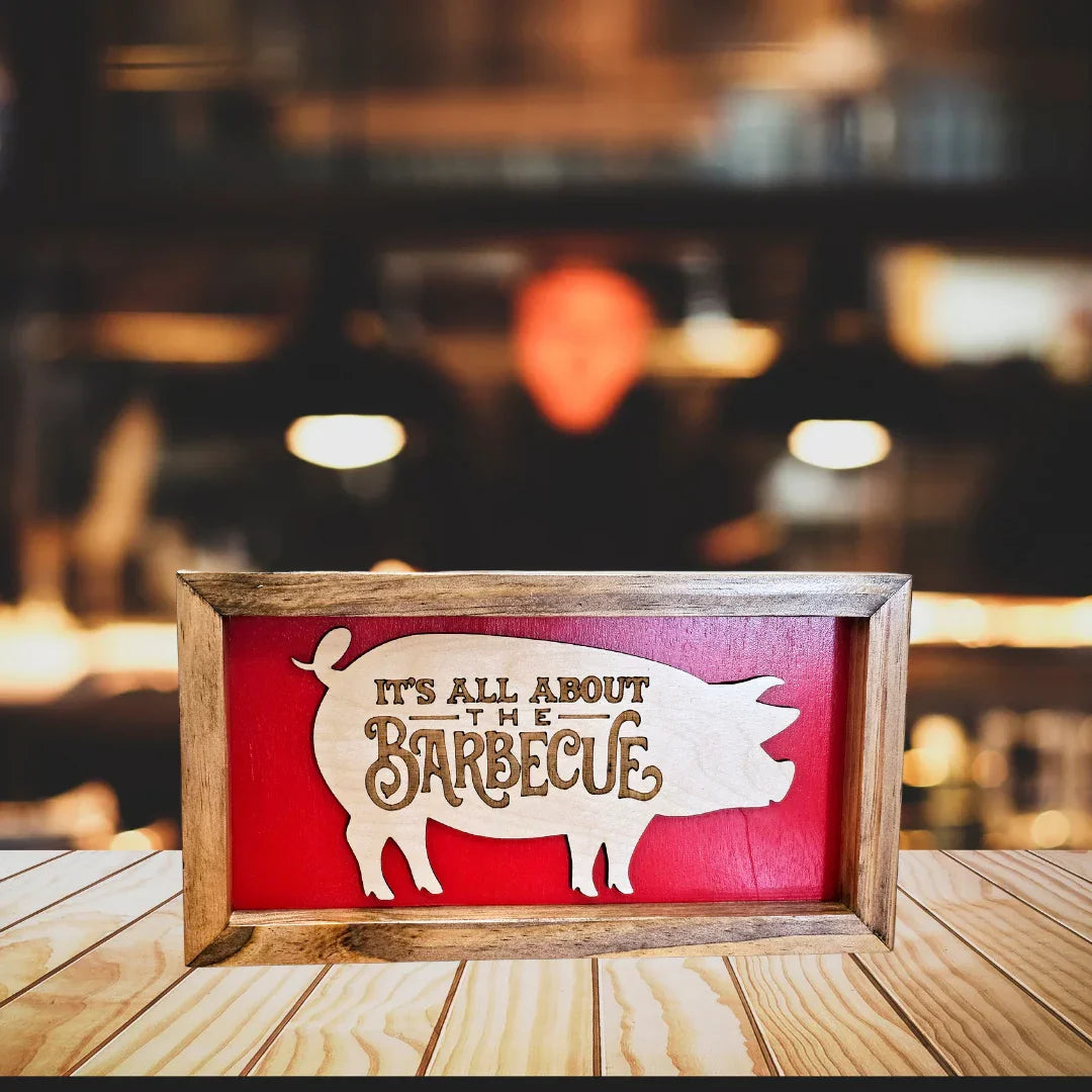 Wooden sign with 'It's All About the Barbecue' text on a red background, blurred restaurant interior in the background.