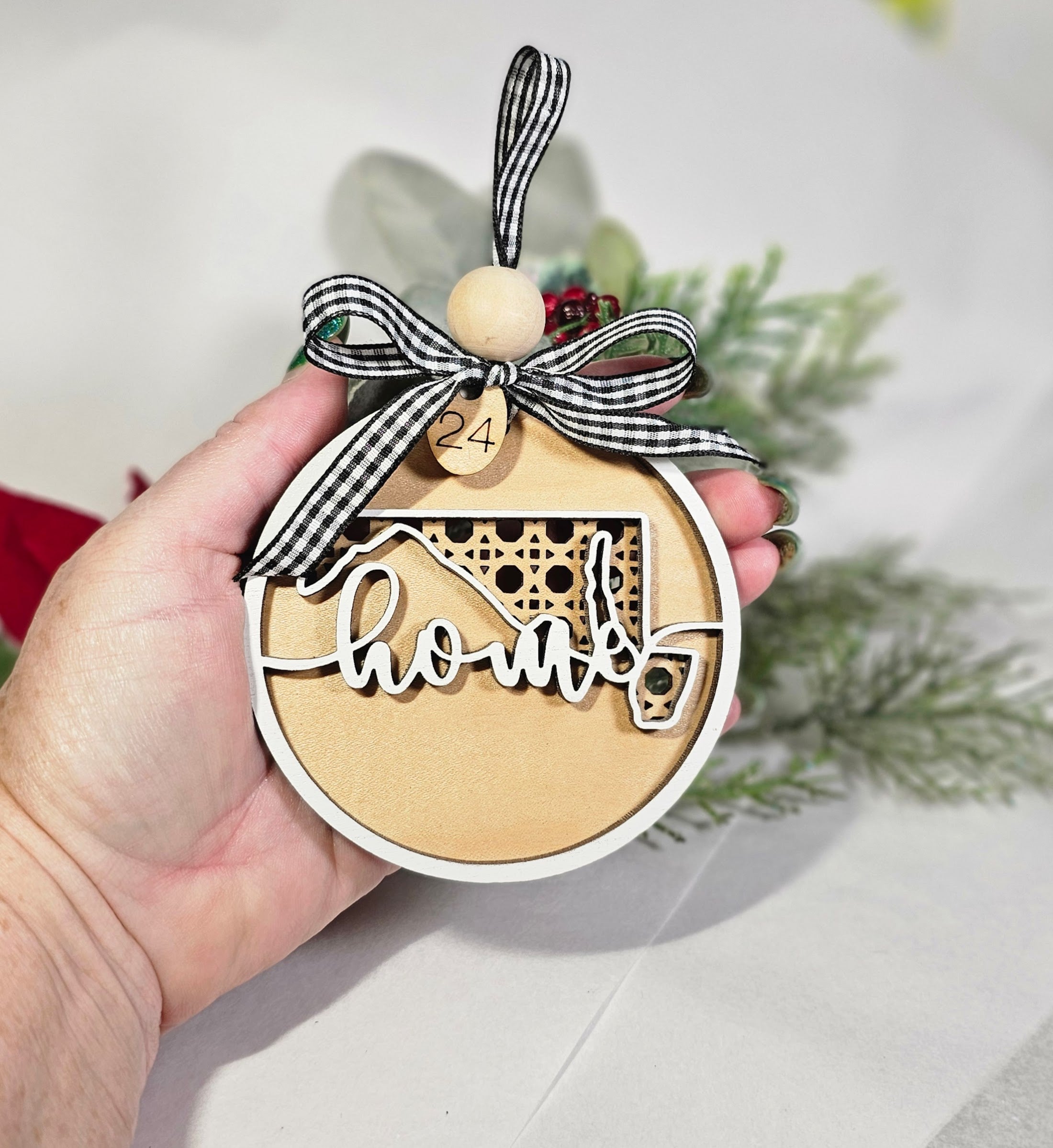 Wooden Maryland ornament with 'home' text and a bow, held by a hand with a blurred background of greenery.