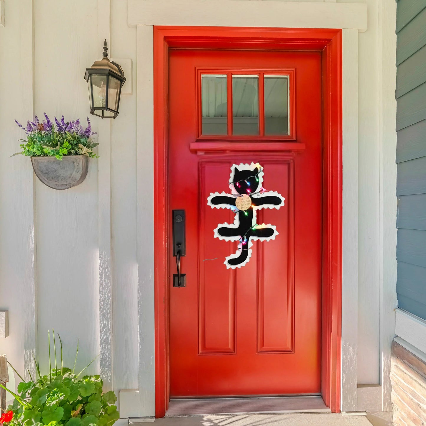 Red front door with a decorative black cat door hanger with electric lights, surrounded by plants and a wall light fixture.
