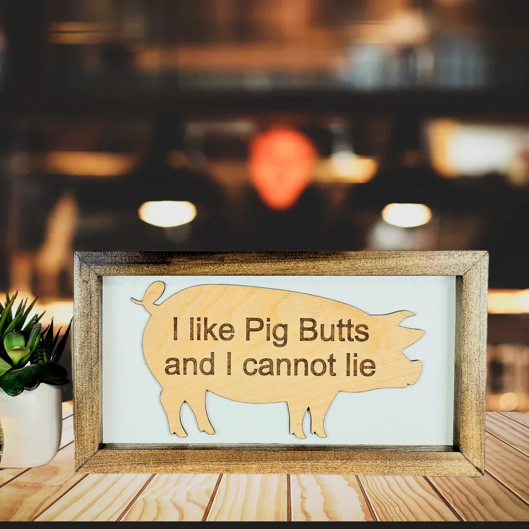 Wooden sign with pig butts illustration and text on a wooden surface with blurred background