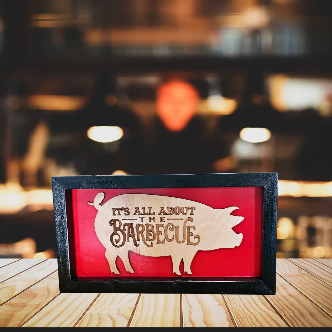 Decorative sign with pig silhouette and 'It's All About the Barbecue' text on a red background, framed in black.