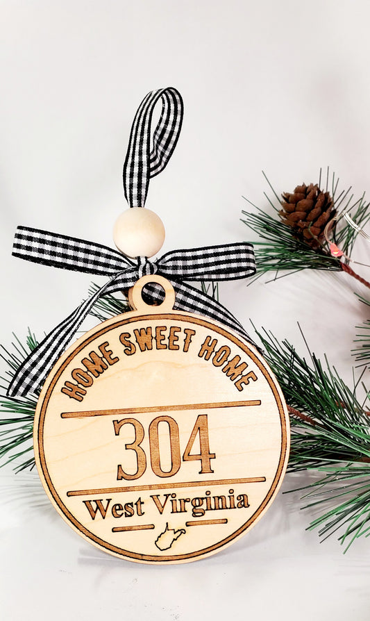 Natural light, birch wood engraved with the phrase, Home Sweet Home along with the area code of 304. West Virginia and the state outline is engraved towards the bottom of the round ornament. A white and black buffalo plaid ribbon and solid, round wood bead adds the finishing touch.