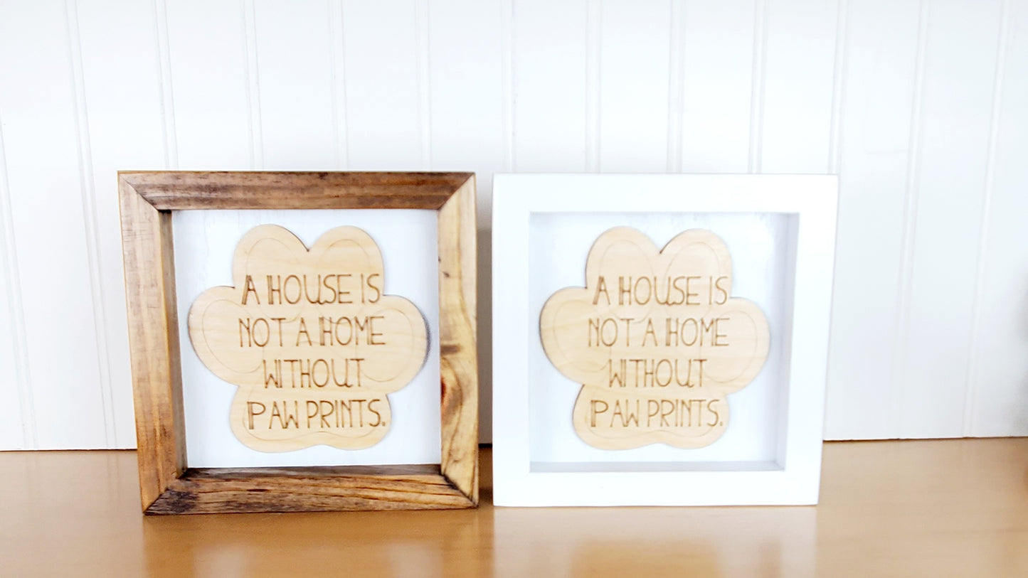 A House is Not a Home Without Paw Prints engraved on natural birch wood cut out in the shape in a paw print and placed on a white background. Choice of walnut stained or white wood frame. 