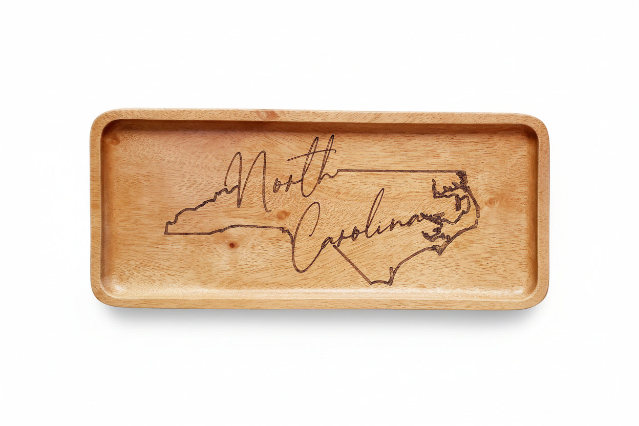 A rectangular wooden platter with an engraved North Carolina outline and non-slip handle grips on the edges, placed on a white surface with decorative white flowers nearby.