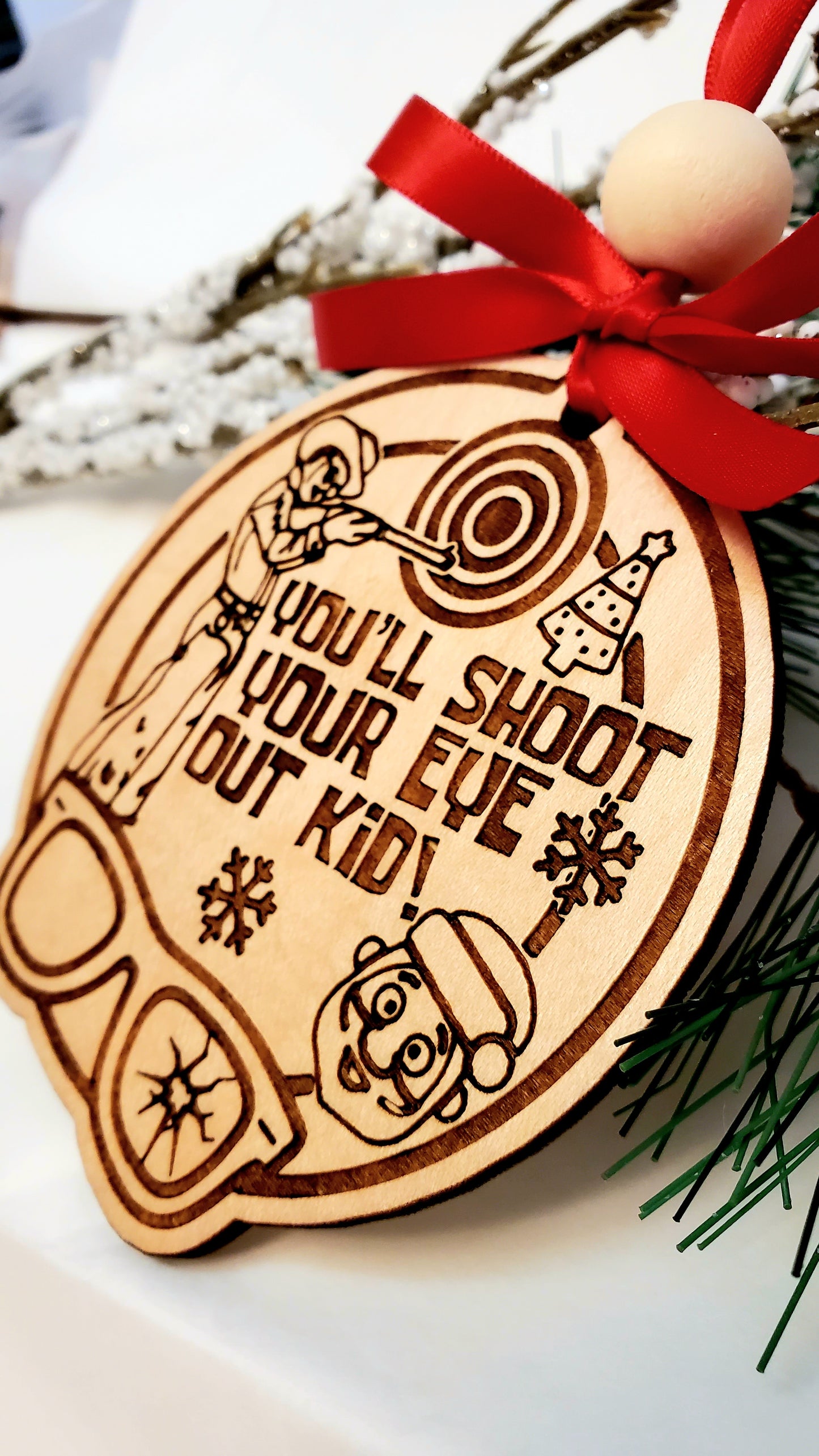 Close-up of You'll Shoot Your Eye Out ornament showing engraved A Christmas Story scene, birch wood with ribbon
