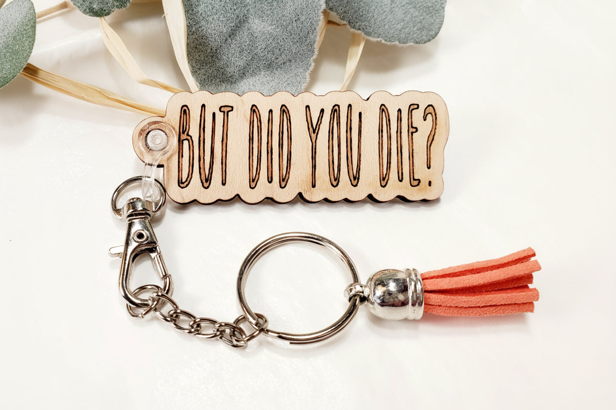 A wooden keychain with a metal ring and a red tassel, featuring the text 'BUT DID YOU DIE?' engraved on it. Funny snarky keychain with humorous saying.