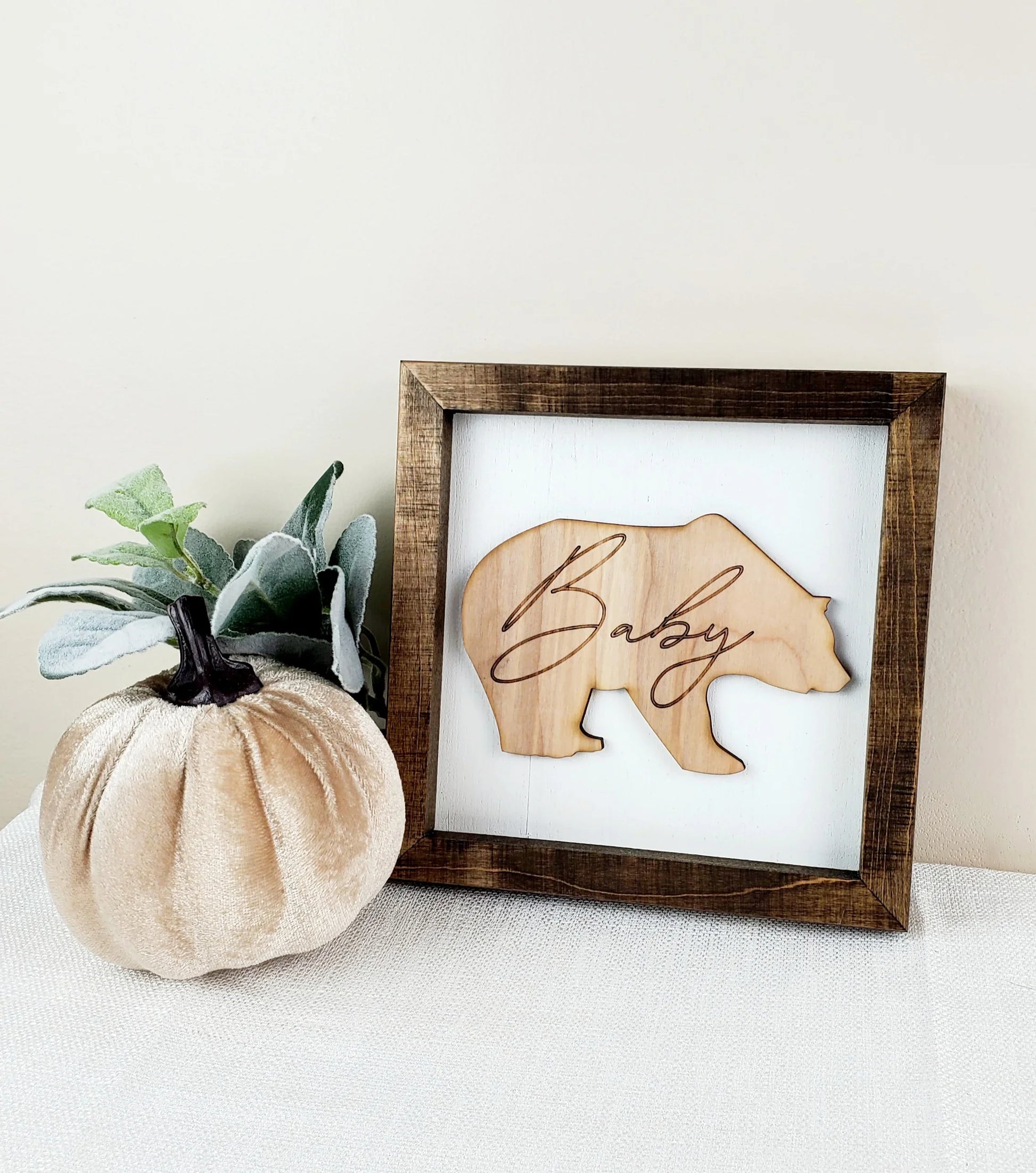 Framed artwork with bear silhouette and 'Baby' text next to a pumpkin and plant on a neutral background