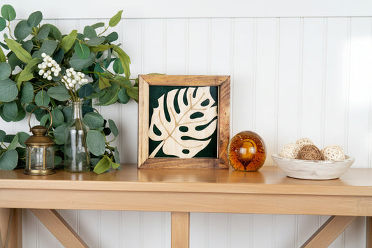 A handmade wooden sign shaped like a monstera leaf with a dark green and natural wood finish, displayed on a white surface with a blurred background of greenery and a decorative glass object to the right.