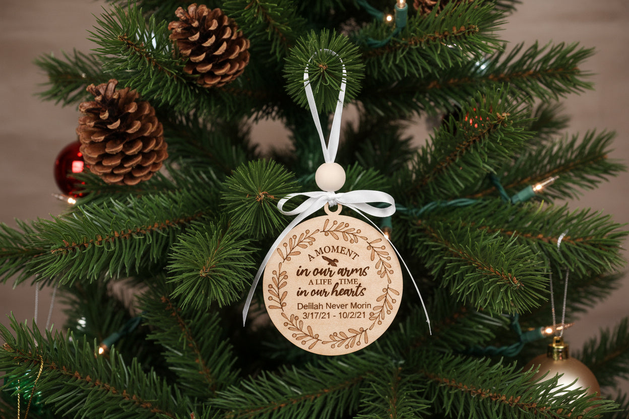 A personalized memorial ornament with a wooden round ornament, white ribbon, and green pine needles, with the inscription 'In our hearts' and birth and death dates.