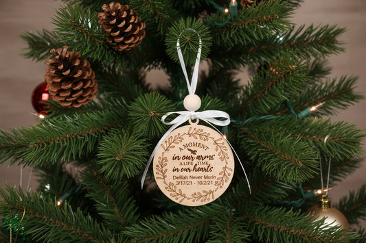A personalized memorial ornament with a wooden round ornament, white ribbon, and green pine needles, with the inscription 'In our hearts' and birth and death dates.