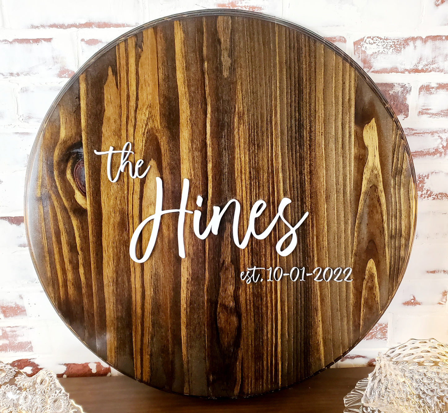 A 2ft round custom wood guest sign board with routered edge and white laser cut names and dates 'The Hines etched on the surface, placed on a brick wall.