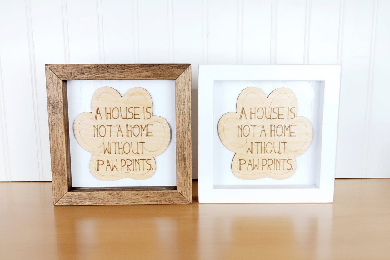 A House is Not a Home Without Paw Prints engraved on natural birch wood cut out in the shape in a paw print and placed on a white background. Choice of special walnut stained or white wood frame. 